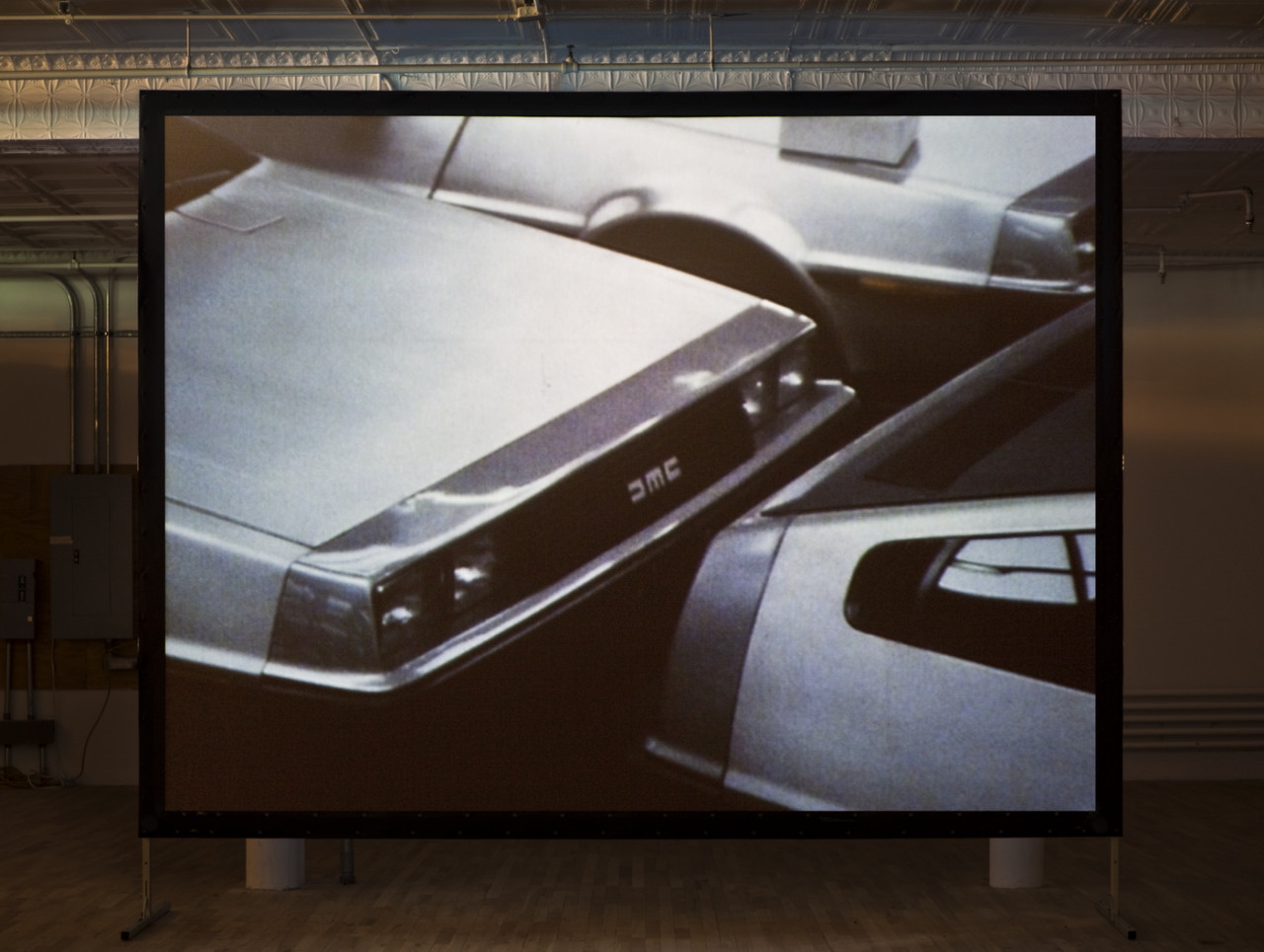 A projected image on a standing screen shows a bumper-to-bumper view of two shiny silver cars with a third car in the background.