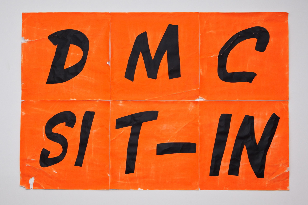Six orange papers arranged in a rectangle are painted with the letters "DMC SIT-IN" in black text.