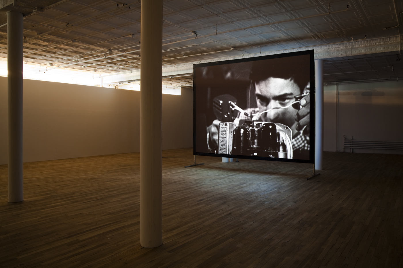 A black and white image of a man working intently on a machine is projected on a standing screen in an open room.