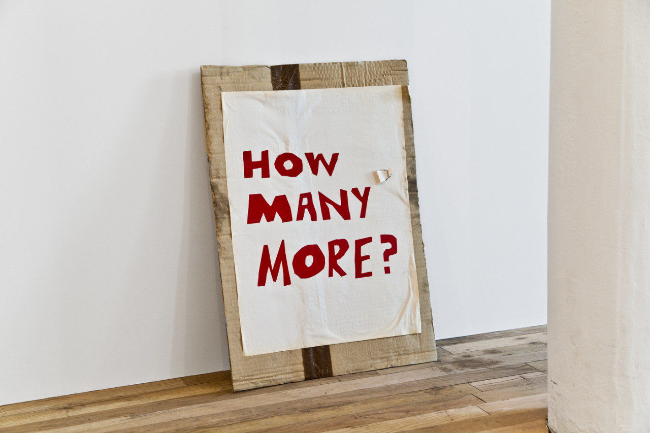 A large piece of paper secured to a piece of cardboard leaning against a wall, slightly scuffed and unfurling from the cardboard. Handwritten, red block text on the paper reads, "HOW MANY MORE?"