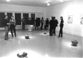 Black and white photograph of people standing in a gallery among five spherical balls on the floor. Framed artworks hang on the right wall.