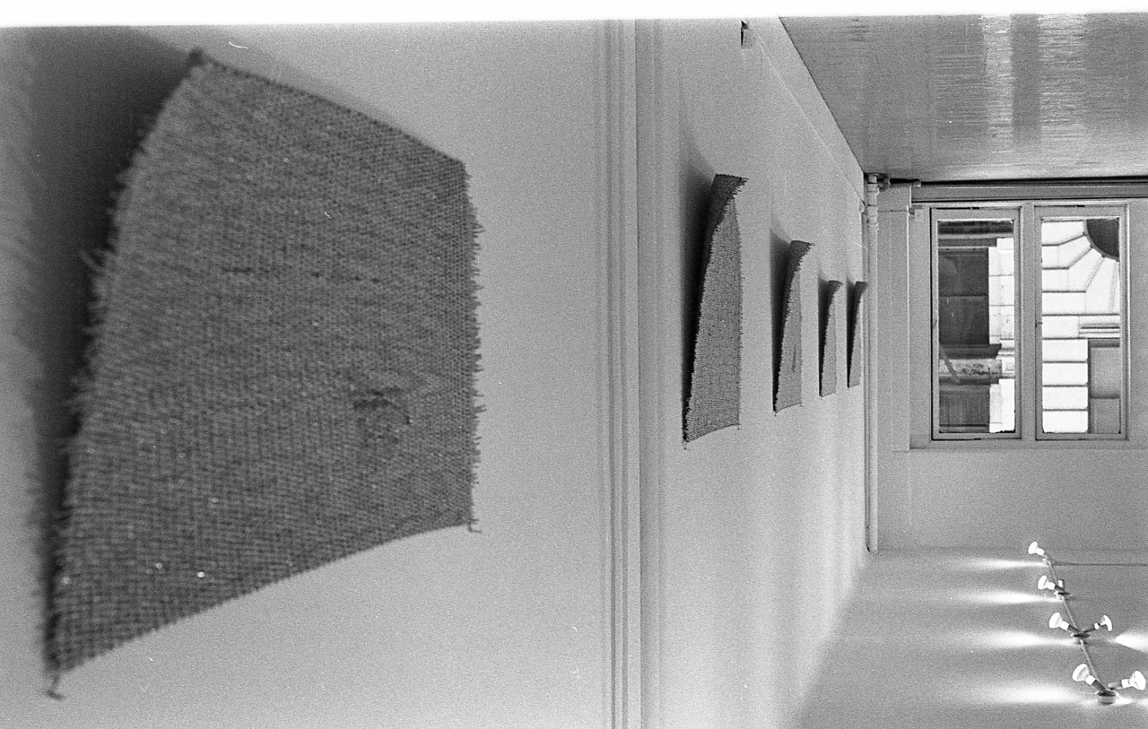 A black and white photograph of a gallery wall. Five mats hang on the wall, their top left corners curving slightly away from the flat surface.