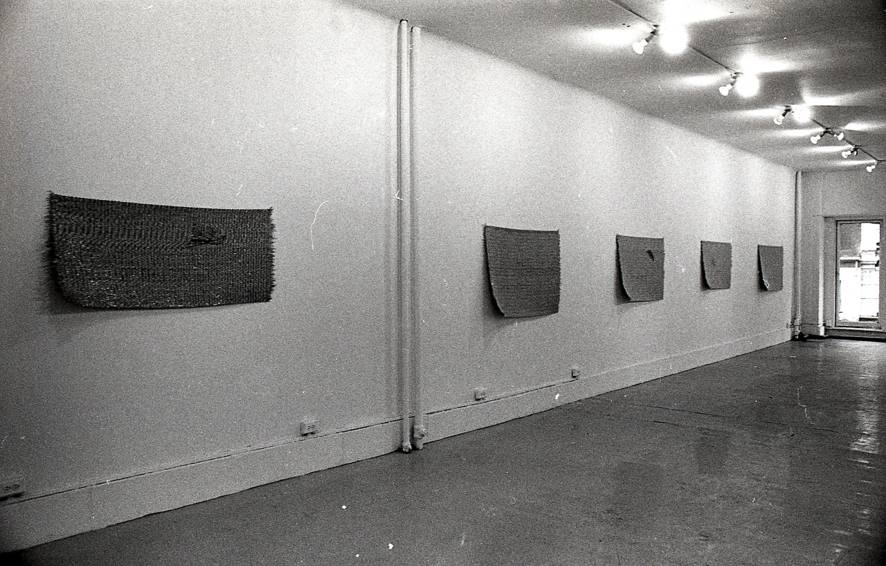 A black and white photograph of a gallery space. Five mats hang on the left wall.