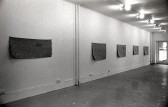 A black and white photograph of a gallery space. Five mats hang on the left wall.