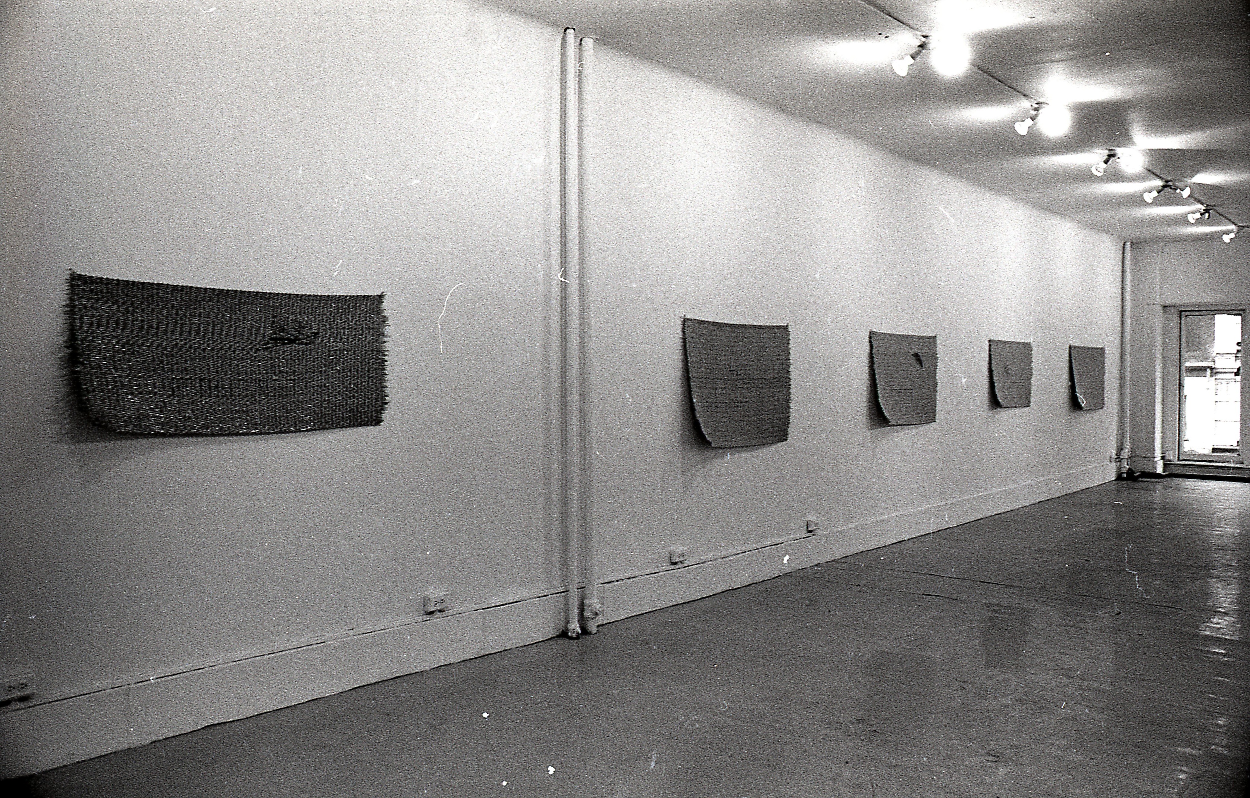 A black and white photograph of a gallery space. Five mats hang on the left wall.
