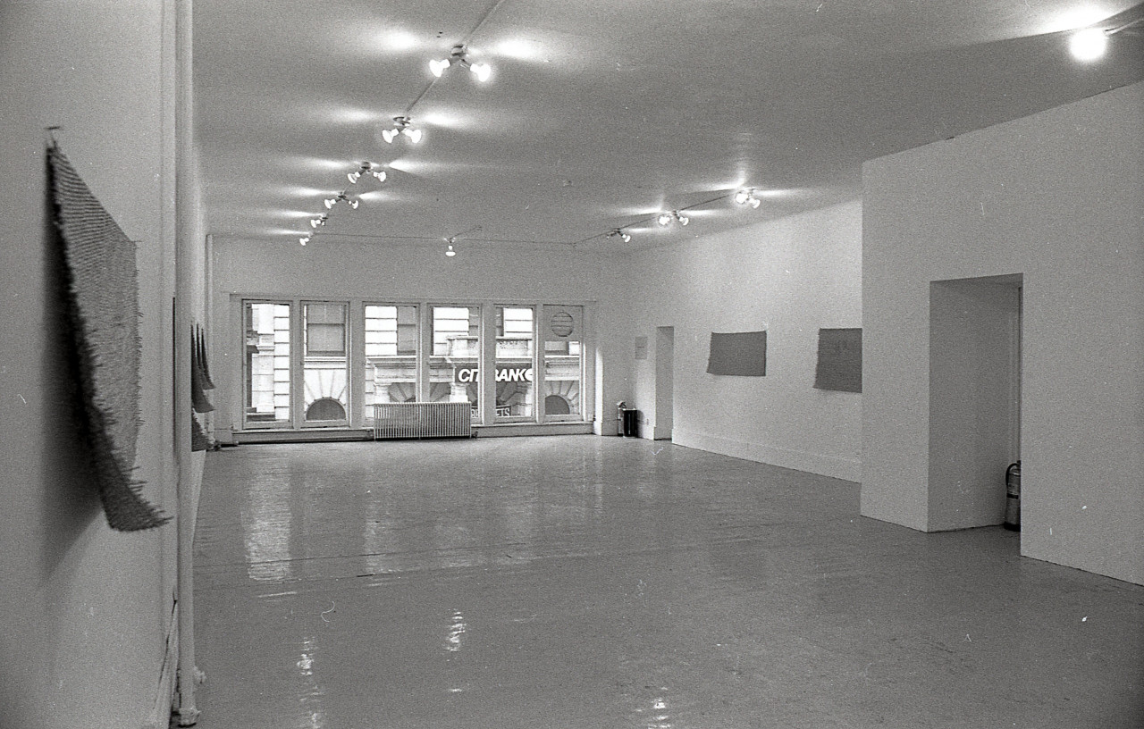 Black and white photograph of a gallery space with windows in the background. Mats hang on the left and right walls.