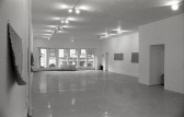 Black and white photograph of a gallery space with windows in the background. Mats hang on the left and right walls.