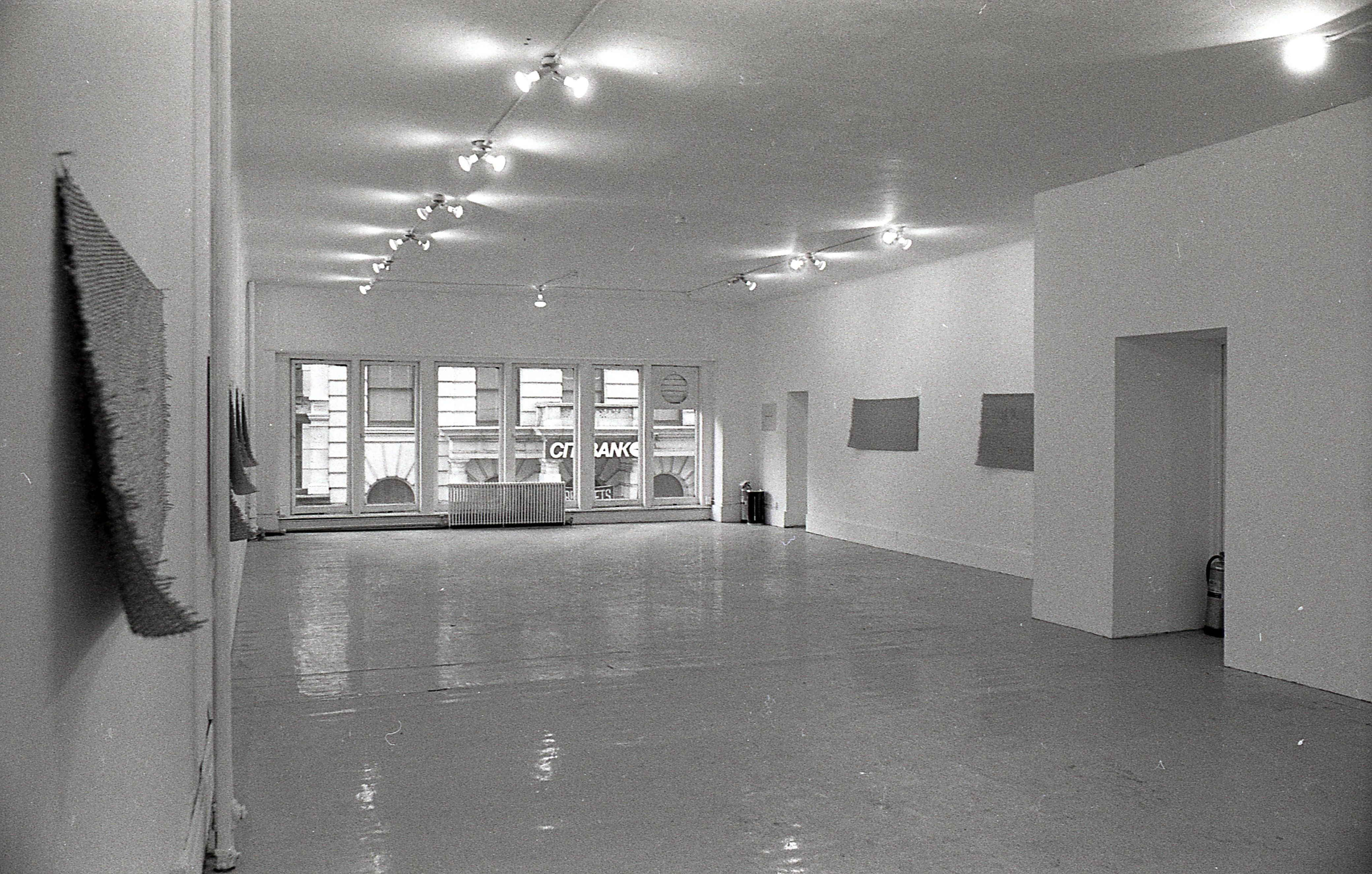 Black and white photograph of a gallery space with windows in the background. Mats hang on the left and right walls.
