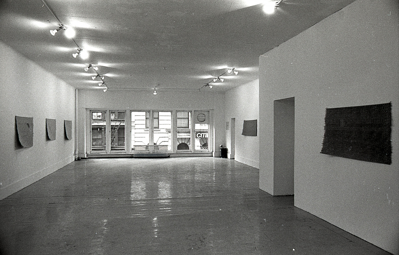 A black and white photograph of a gallery space with windows in the background. Mats hang on the left and right walls.