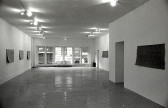 A black and white photograph of a gallery space with windows in the background. Mats hang on the left and right walls.