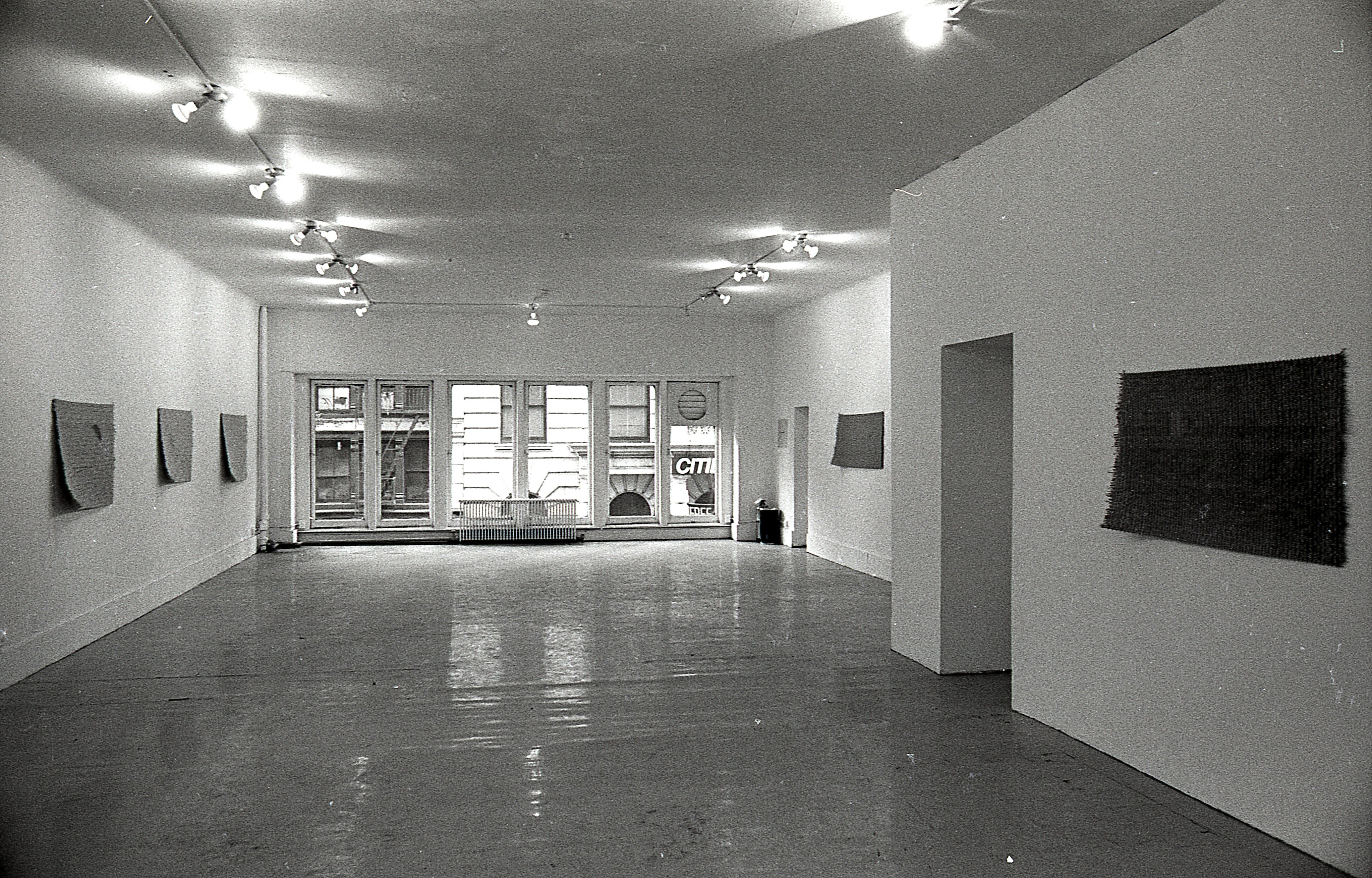 A black and white photograph of a gallery space with windows in the background. Mats hang on the left and right walls.