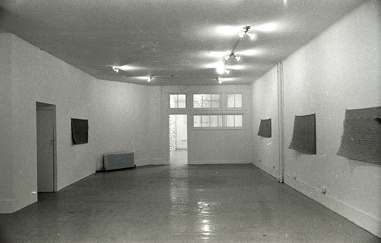 A black and white photograph of a gallery space with windows and an open door in the background. Four mats hang on the walls.