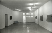 A black and white photograph of a gallery space with windows and an open door in the background. Four mats hang on the walls.