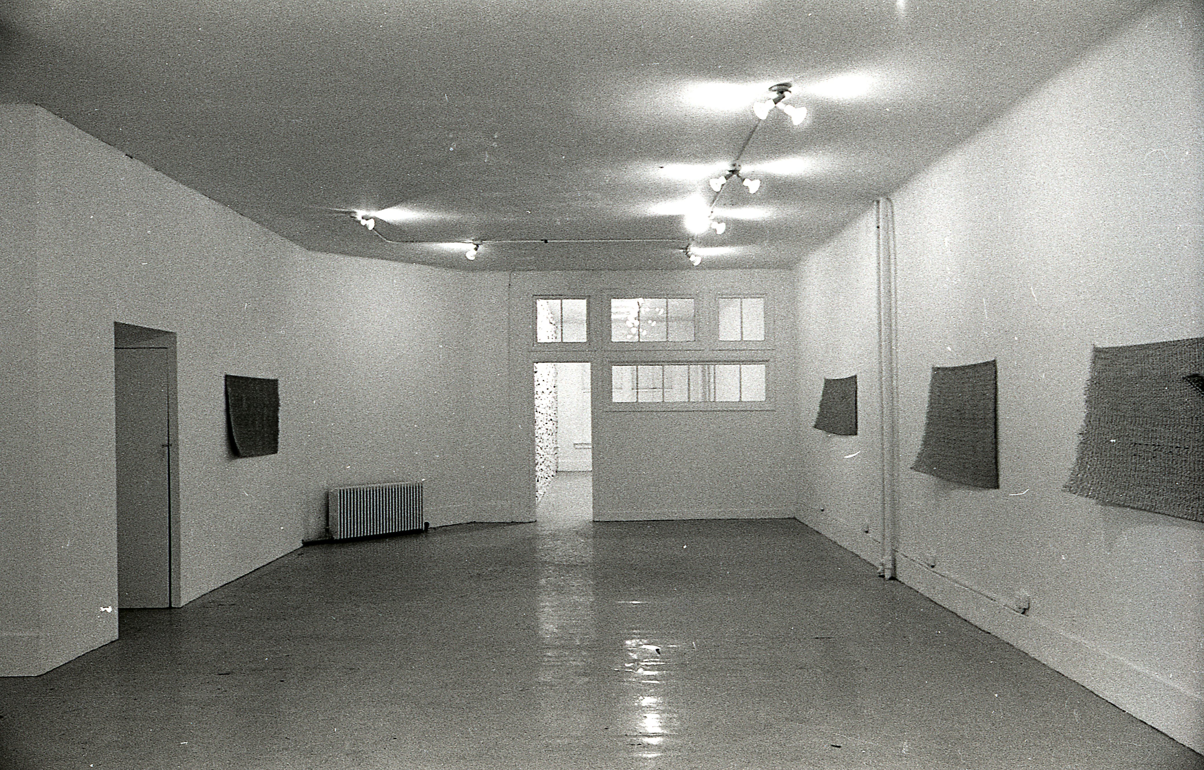 A black and white photograph of a gallery space with windows and an open door in the background. Four mats hang on the walls.