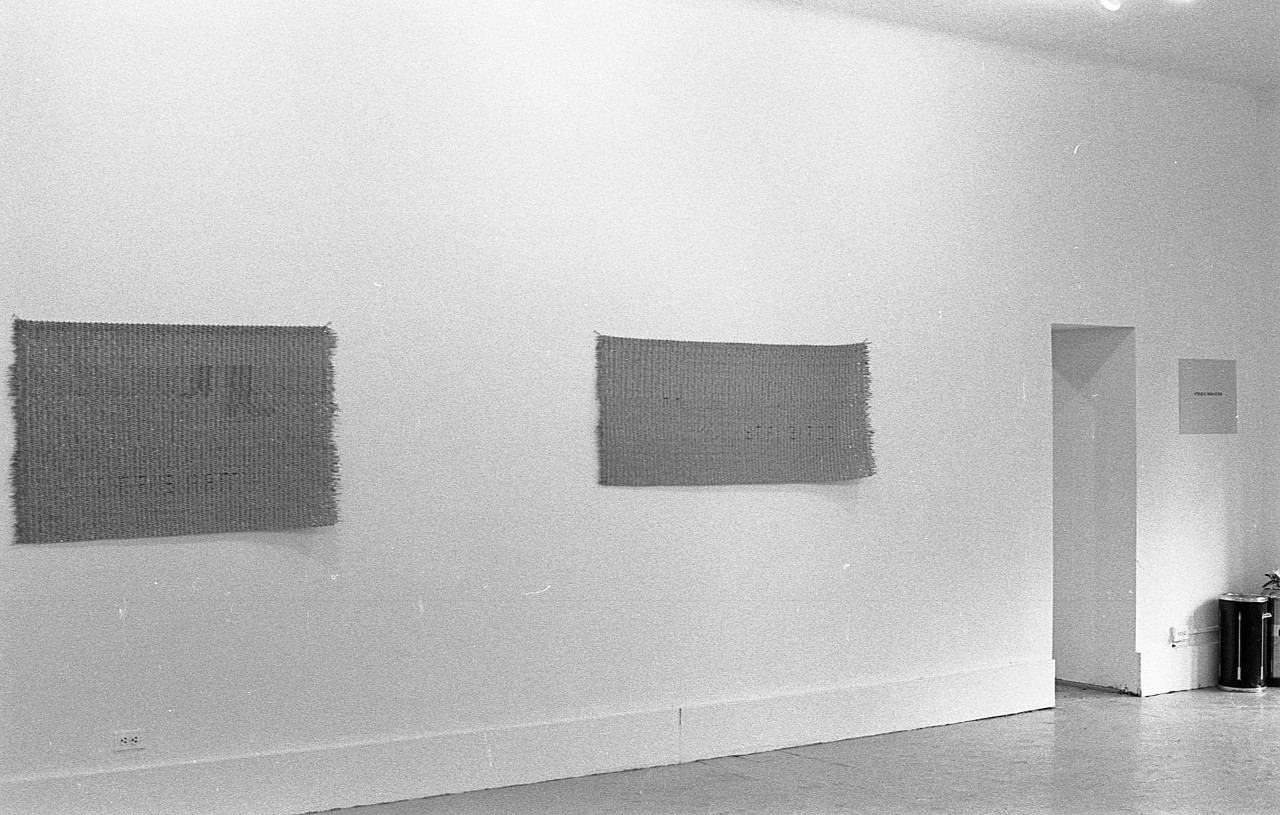 A black and white photograph of a gallery wall, two hanging mats and a doorframe are visible.