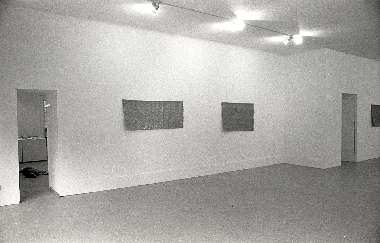 A black and white photograph of a gallery space, three mats are visible hanging on the wall.