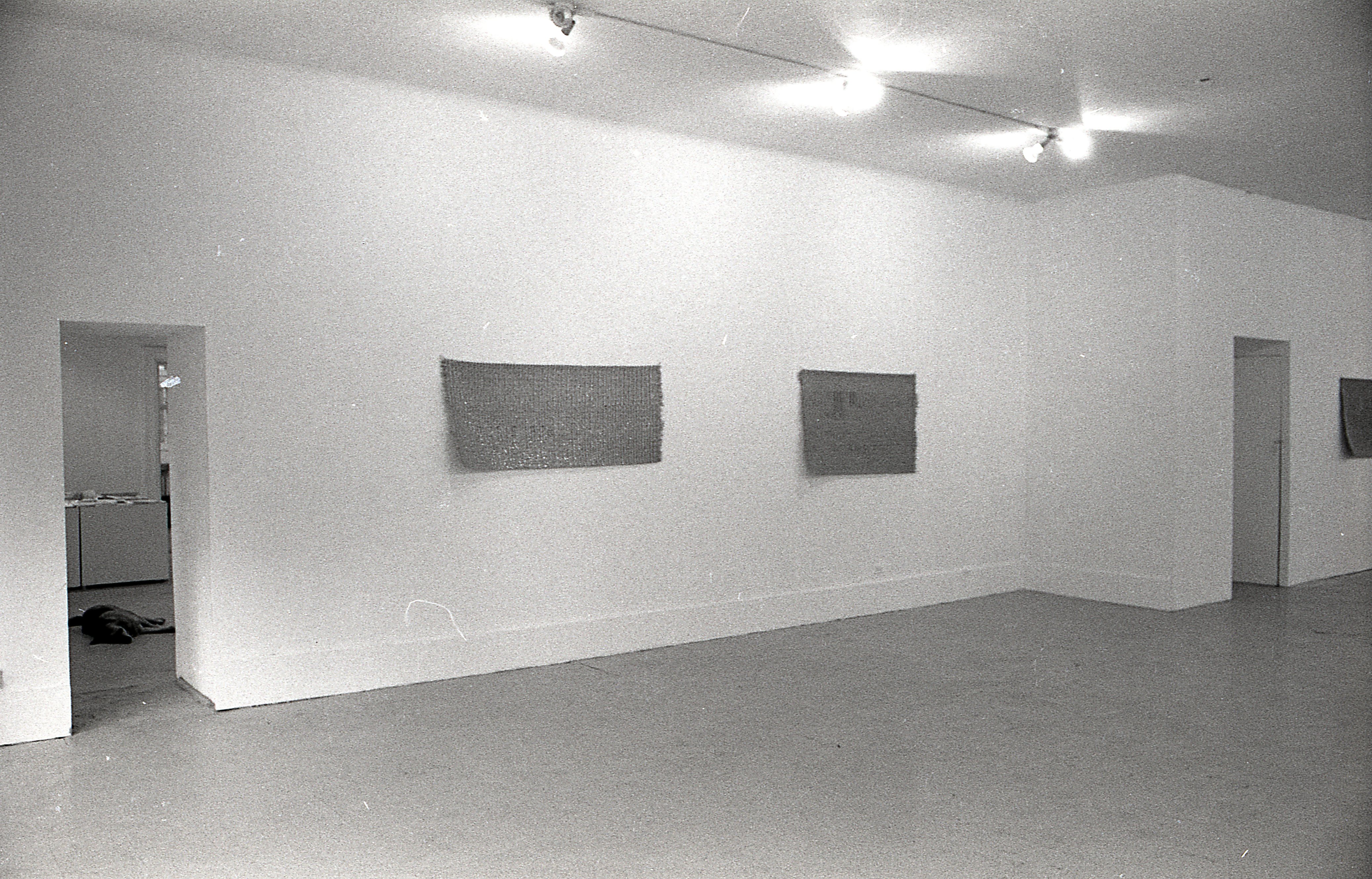 A black and white photograph of a gallery space, three mats are visible hanging on the wall.