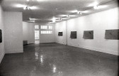 A black and white photograph of a gallery space with windows and an open door in the background. On the right, four mats hang on the wall.