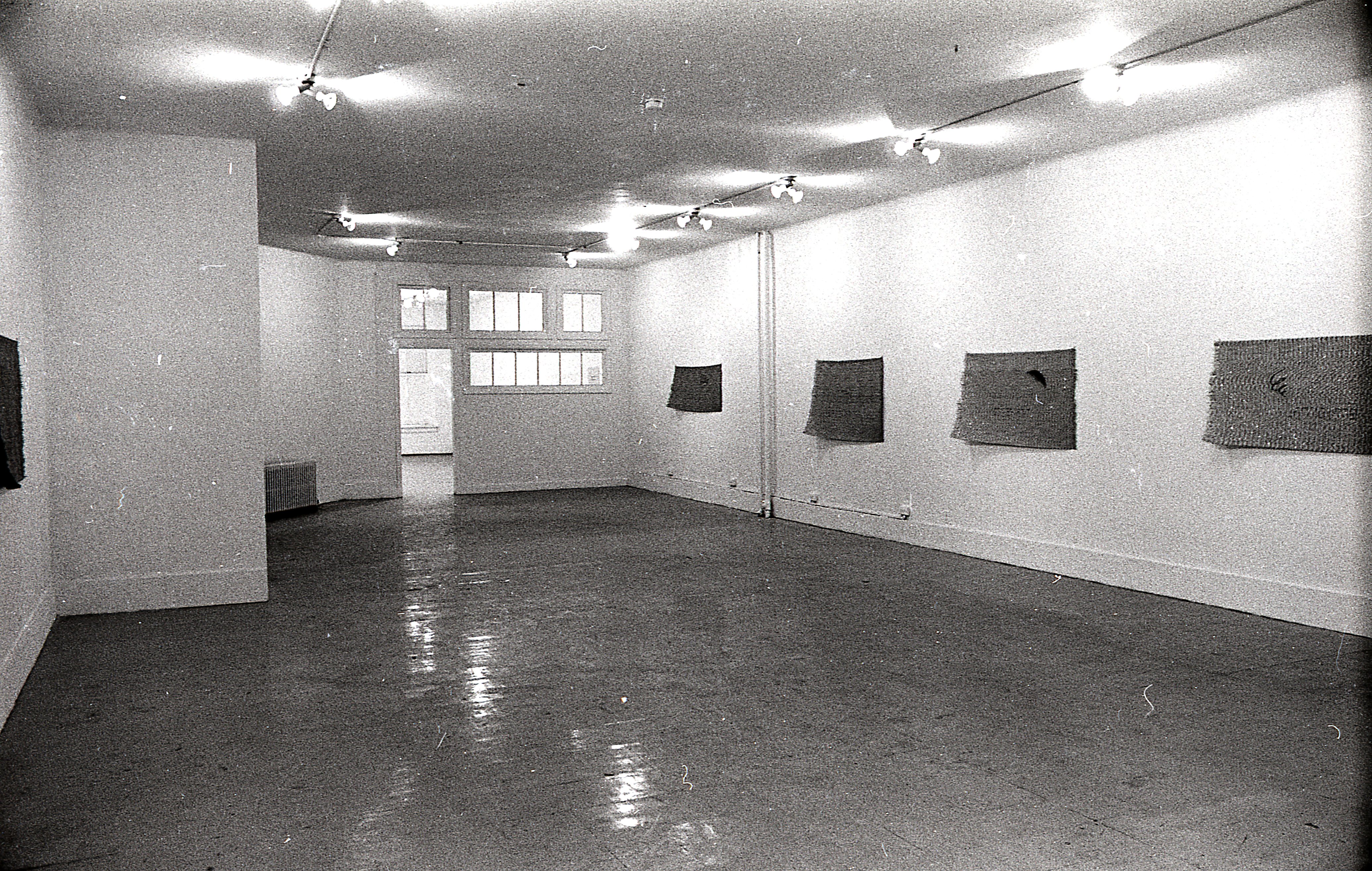 A black and white photograph of a gallery space with windows and an open door in the background. On the right, four mats hang on the wall.