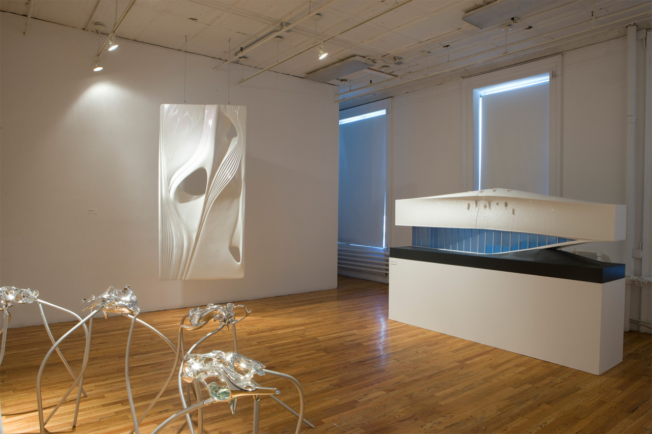 Several sculptures and models sit in a gallery space with wooden floors. To the left of the image, there are several shining metalic forms supported by long, spider-like legs standing at hip height. On the image