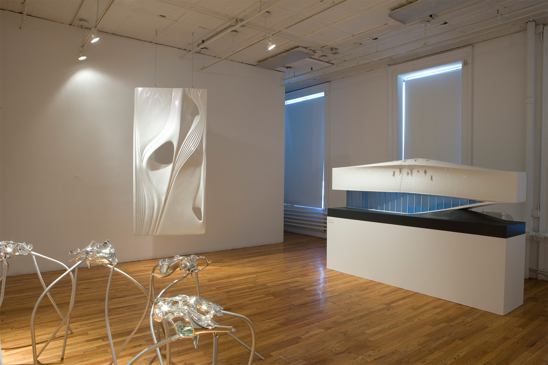 Several sculptures and models sit in a gallery space with wooden floors. To the left of the image, there are several shining metalic forms supported by long, spider-like legs standing at hip height. On the image's right is a large model of a futuristic building. Hanging from the ceiling behind the metal sculptures is a shiny, white rectangular sculpture with sleek, ribbon-like forms.