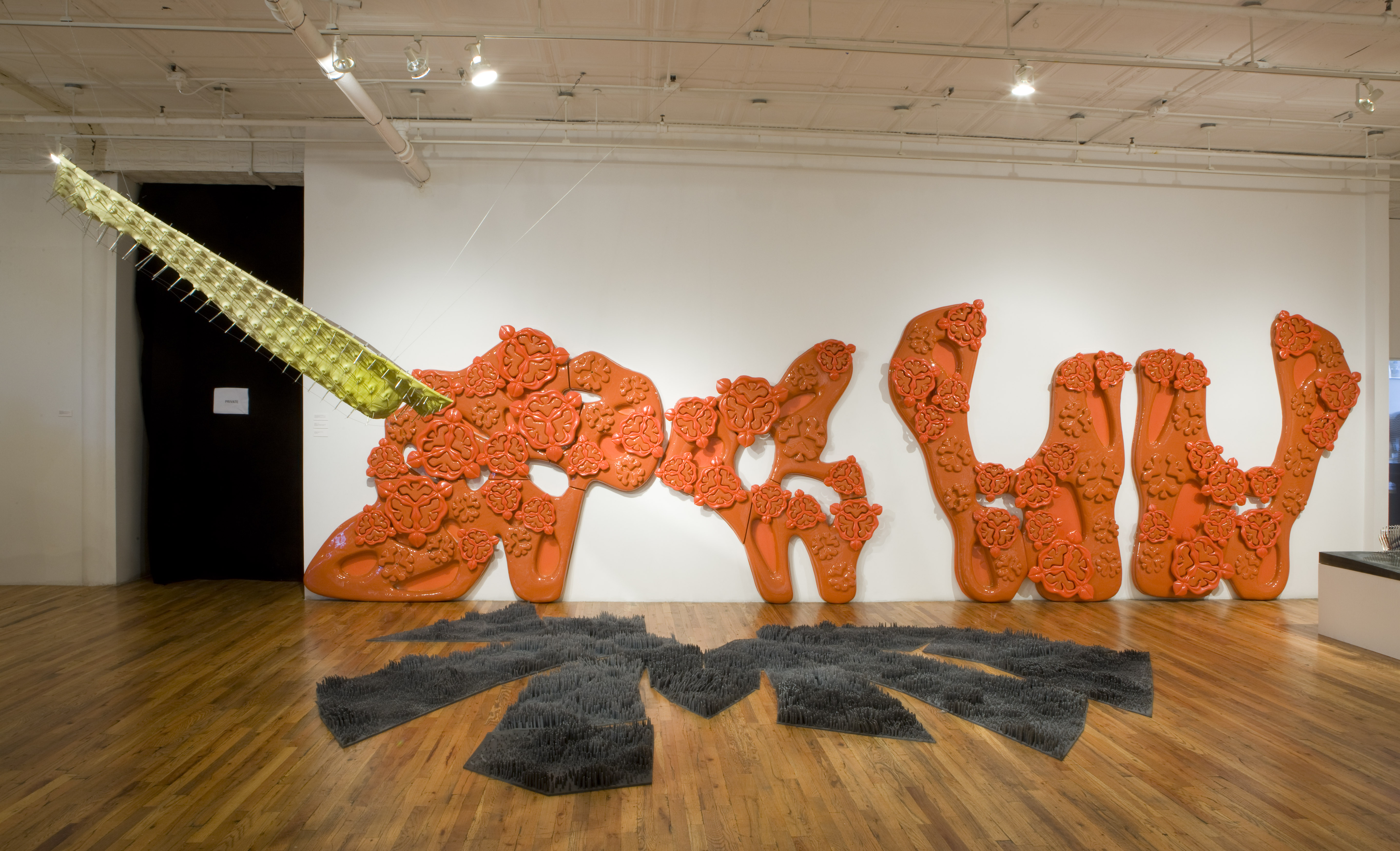 Three sculptural forms sit and hang in a large gallery space. A large gray form with anemone-like tendrils is spread across the floor. Behind it on the gallery wall there is a large, shiny orange sculpture covered in bud-like forms. To the left of the space hangs a yellow sculpture with metal spikes protruding from its surface.