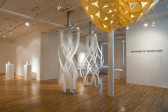 Two large structures made of long, white strips that swirl around one another and form a vertical column are mounted from the ceiling and floor of an open gallery space. Black text posted on a gallery wall to the right of the image reads, 
