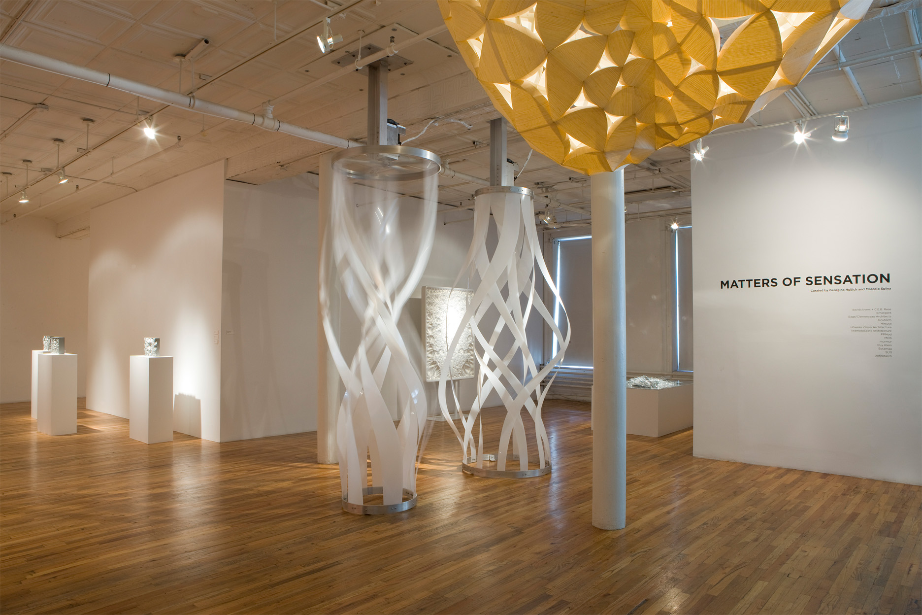 Two large structures made of long, white strips that swirl around one another and form a vertical column are mounted from the ceiling and floor of an open gallery space. Black text posted on a gallery wall to the right of the image reads, 