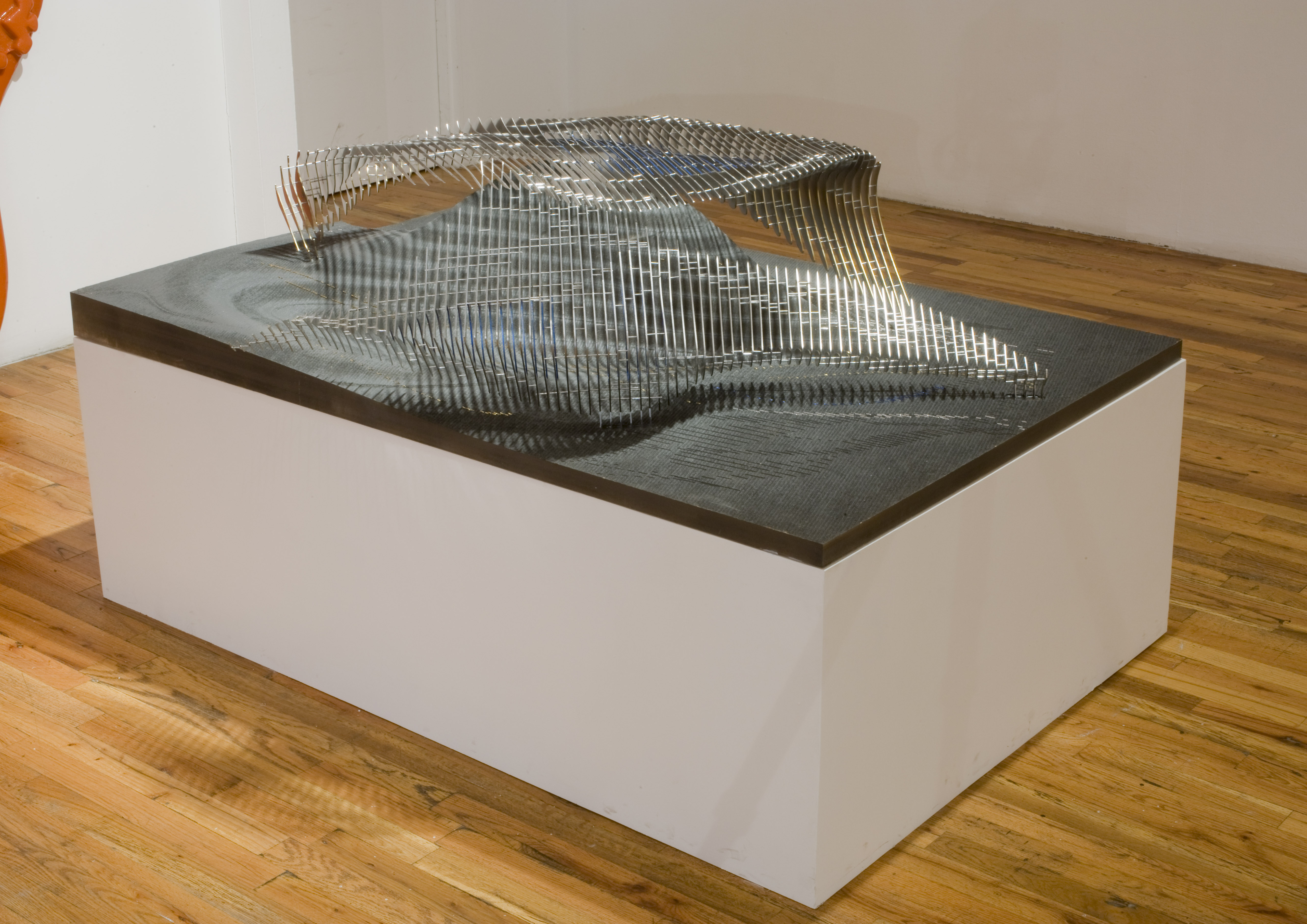 A metal form sits on a sloped gray slap on top of a low, rectangular plinth. The 3D form is composed of thin metal sheets that together form an amorphous shape that casts a series of shadows on the surface below it.