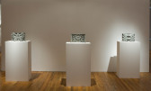 Three gray objects rest on separate rectangular, white plinths in a gallery space with wooden flooring. Each of the objects is rectangular and has a 3D, biomorphic form.
