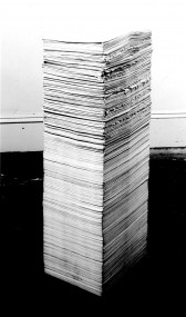 A black and white installation photograph of a tall stack of papers in a gallery space.