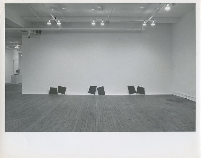 A black and white installation photograph of a gallery space. Placed on the floor pairs of dark squares lean against a white wall on the far end.