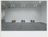 A black and white installation photograph of a gallery space. Placed on the floor pairs of dark squares lean against a white wall on the far end.