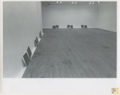 A black and white installation photograph of a gallery space. Placed on the floor, pairs of dark squares lean against a white wall.