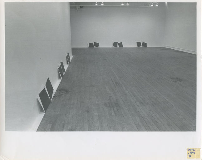 A black and white installation photograph of a gallery space. Placed on the floor, pairs of dark squares lean against a white wall.