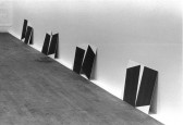 A black and white installation photograph of a gallery space. Placed on the floor, canvasses of dark rectangles on white backgrounds lean against a white wall.