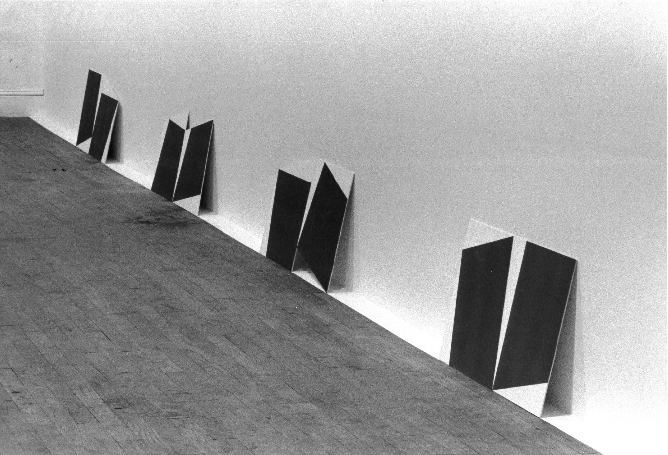 A black and white installation photograph of a gallery space. Placed on the floor, canvasses of dark rectangles on white backgrounds lean against a white wall.