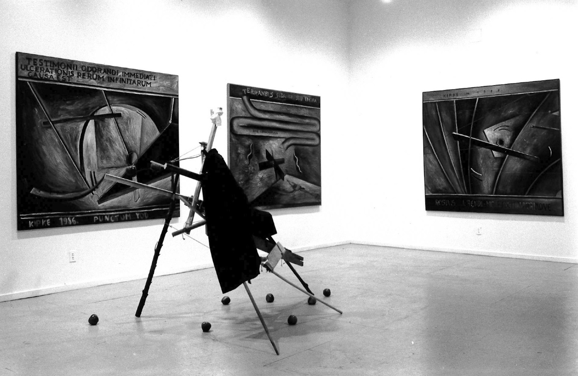 An abstract sculpture made of sticks, cords, and a textile sits in the center of a gallery space.  Three large paintings hang on the walls around it, all blending abstract, painterly designs with latin text.