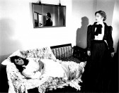 A black and white film still of two people in period dress.  One lies on a patterned couch, wrapped in sheets, while the other stands observing.  Above the couch, a mirror on the wall reflects the scene to the camera.