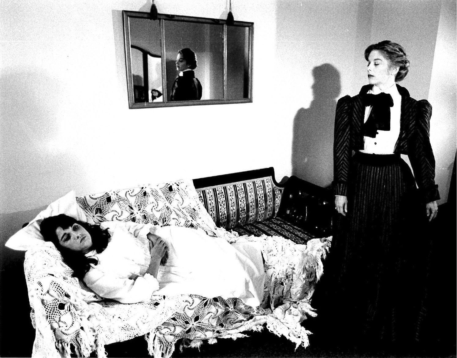 A black and white film still of two people in period dress.  One lies on a patterned couch, wrapped in sheets, while the other stands observing.  Above the couch, a mirror on the wall reflects the scene to the camera.