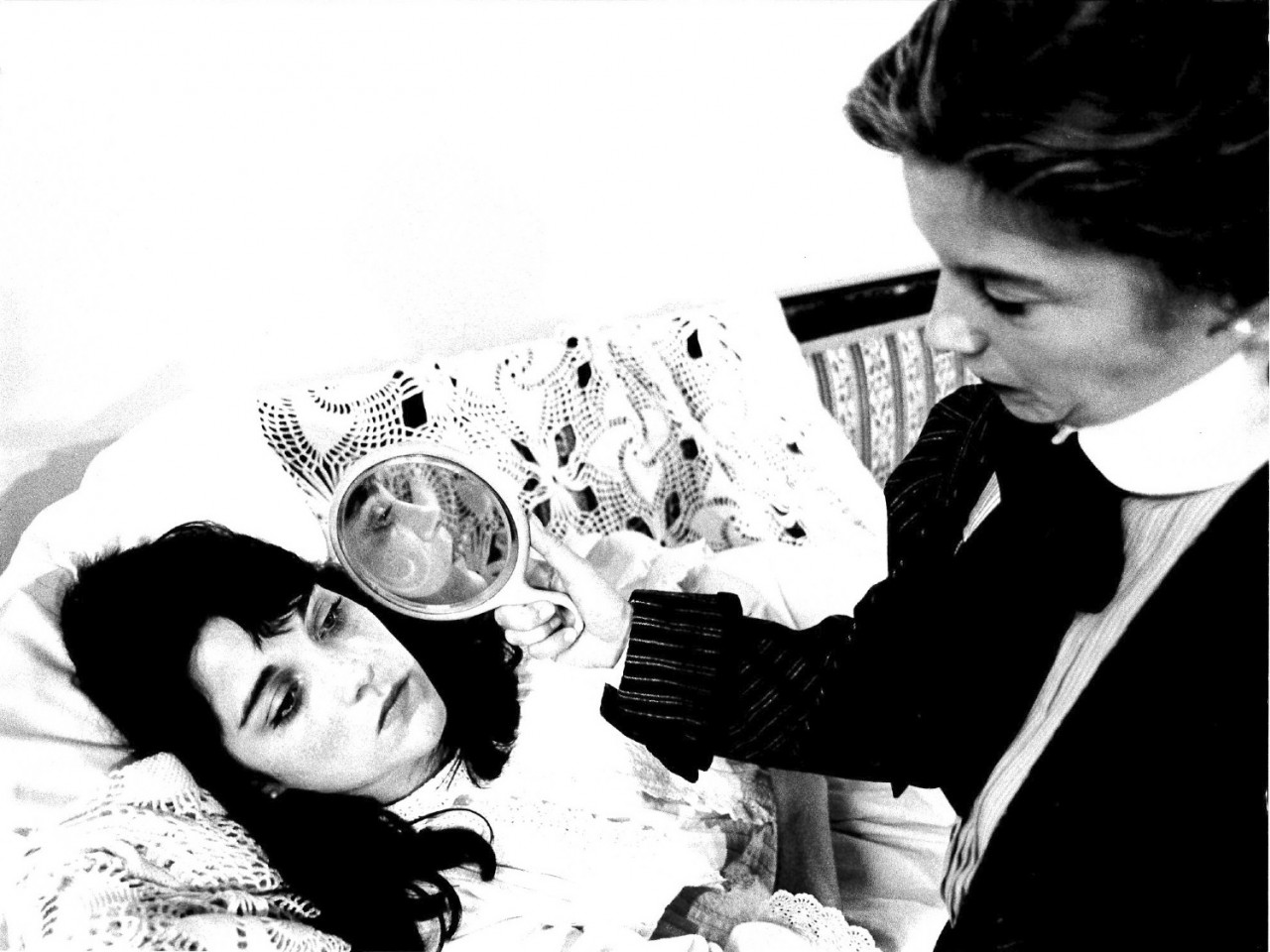 A black and white film still of a person lying against embroidered pillows.  Another person sits alongside her and holds a mirror up to her face, reflecting it towards the camera.