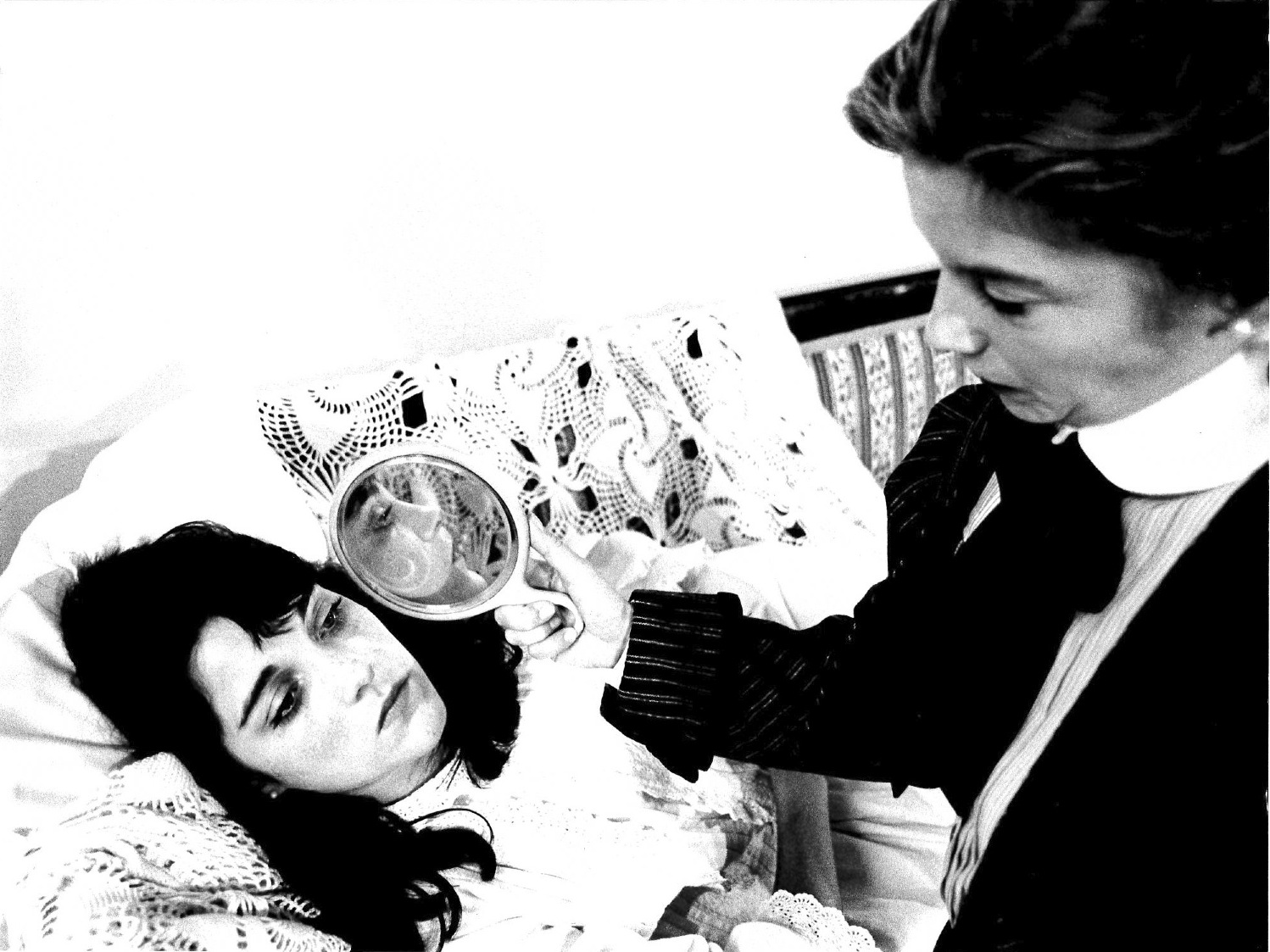 A black and white film still of a person lying against embroidered pillows.  Another person sits alongside her and holds a mirror up to her face, reflecting it towards the camera.