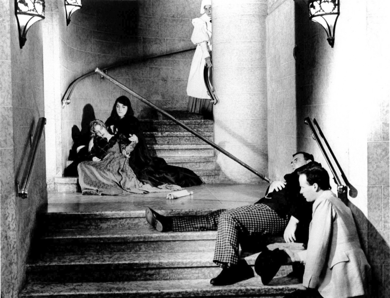 A black and white film still of four people in period dress sprawled on a curving stone stairwell.  Two of these people appear to have fainted, and are supported by the other two.  At the top of the stairs, a person in a maid