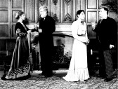 A black and white film still of four people in period dress standing in from of an ornately carved fireplace.  The people stand in two pairs, apparently engaged in conversation.