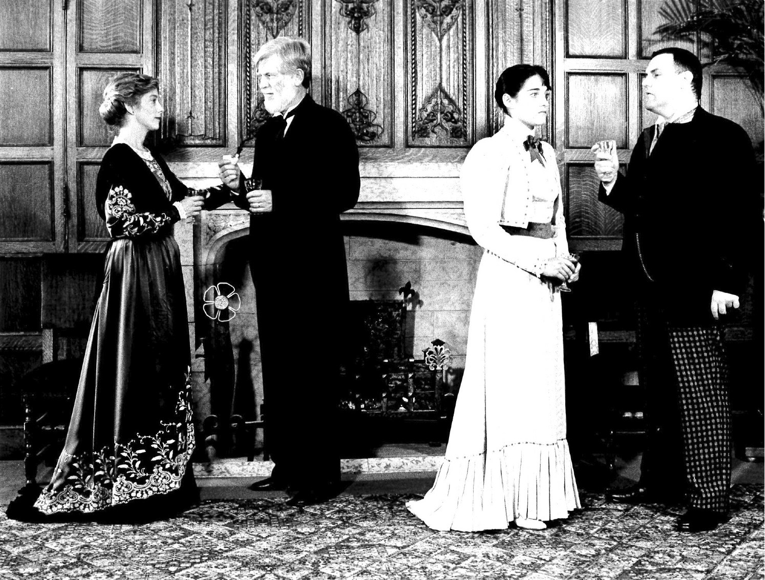A black and white film still of four people in period dress standing in from of an ornately carved fireplace.  The people stand in two pairs, apparently engaged in conversation.
