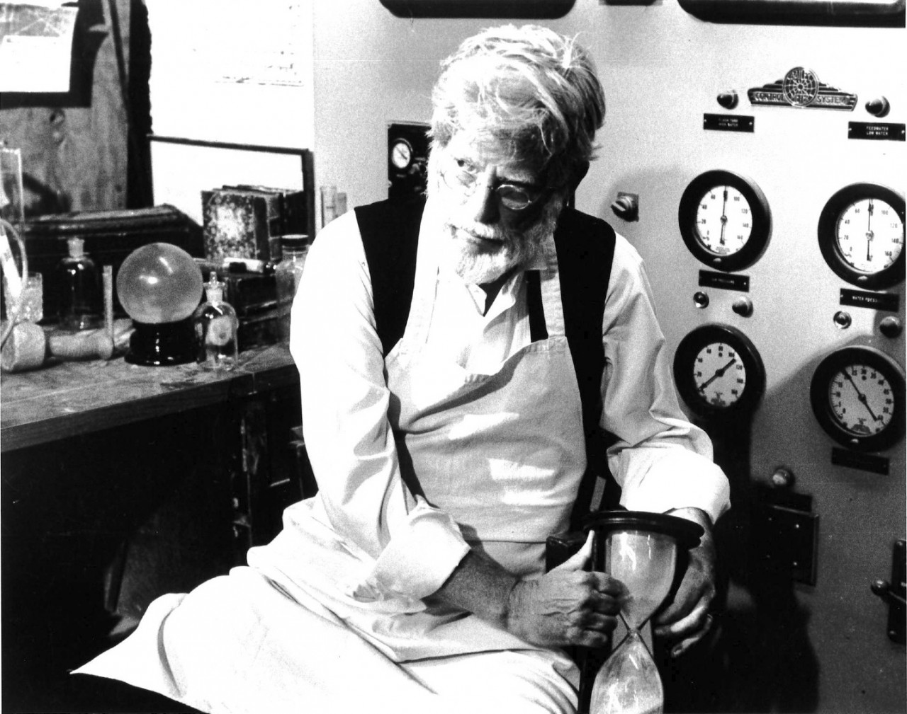 A black and white film still of a person with a white beard wearing an apron seated in front of a workbench covered in glass bottles and scientific instruments.  He holds an hourglass and the wall behind him is lined with dials.