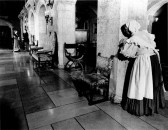 A black and white film still of two people in period dress engaged in conversation at the far end of an ornate allway lit by candlelight.  In the foreground, a person in a maid's costume peeks around one end of a colonnade, observing.