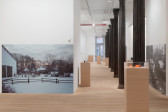 A large gallery space houses a variety of plinths made from MDF, atop of which are various archival objects. On the walls are large photographs affixed directly to the wall.