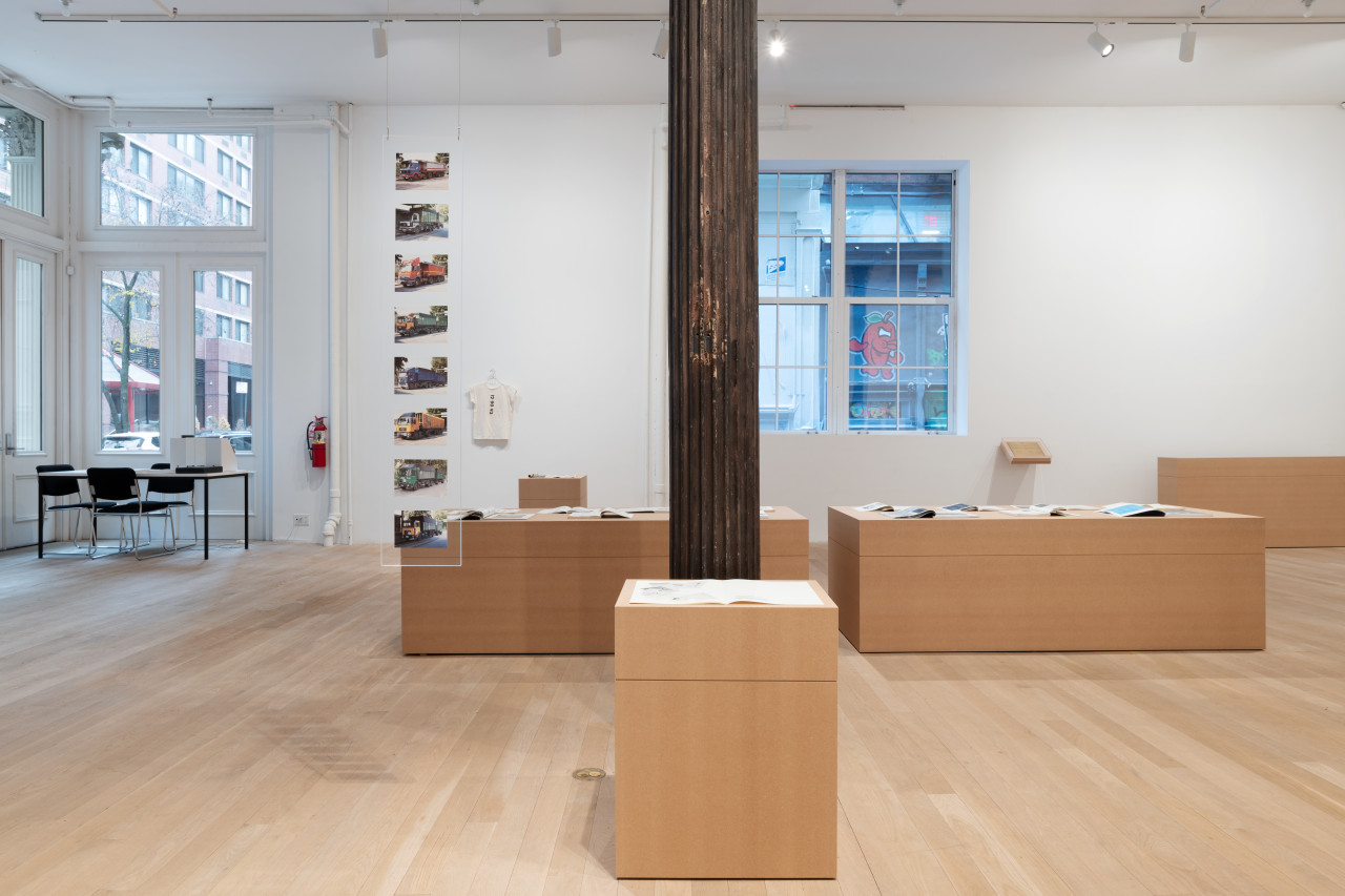 A large gallery space houses a variety of plinths made of MDF atop of which are placed various archival documents and objects, such as exhibition pamphlets and art historical journals.