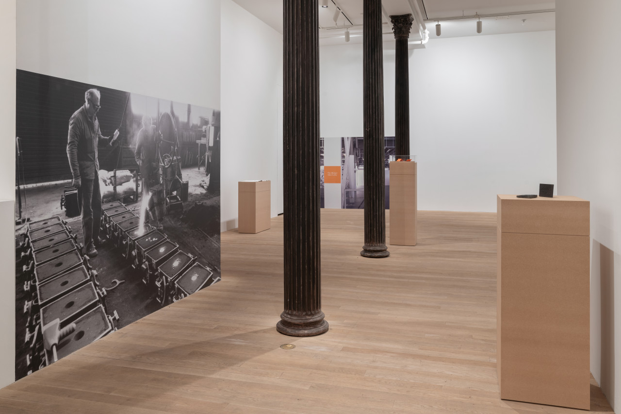 The hallway of a gallery, leading into a large gallery space. On the left wall is a large black-and-white photograph is affixed to the wall, depicting the process of casting the iron objects displayed on the opposite wall on a tall plinth made of MDF. In the room in the back ground are more plinths and archival objects and documentation.